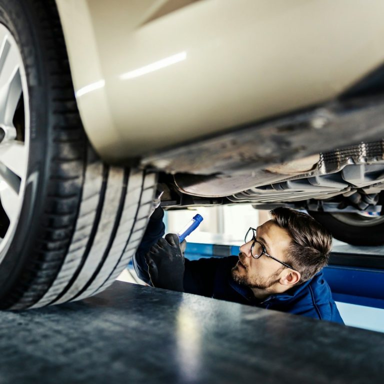 Mechaniker überprüft das Auto von unten, trägt eine Brille und nutzt Werkzeug.