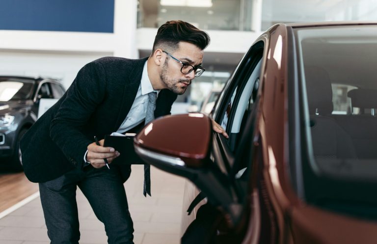 Ein Mann in Anzug betrachtet aufmerksam den Seitenspiegel eines roten Autos im Autohaus.